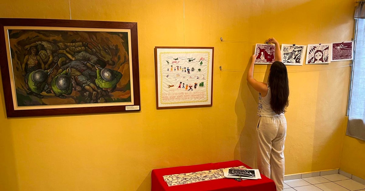 A yellow wall in a museum covered in artwork with a woman in pale clothing hanging pictures. Paintings on the wall illustrate atrocities.