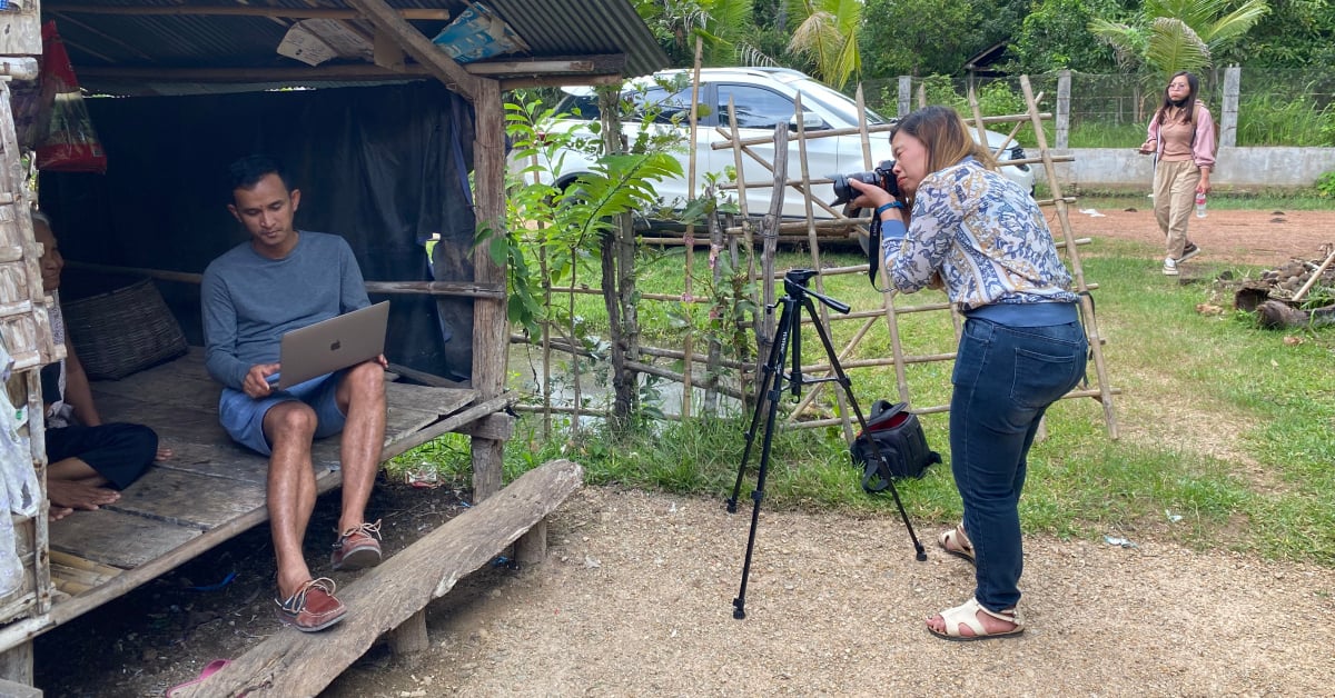 On the left, two people (one obscured) sit in an open hut and look at something on a laptop screen. On the right, a woman stands in front of a tripod and takes a photograph of them.