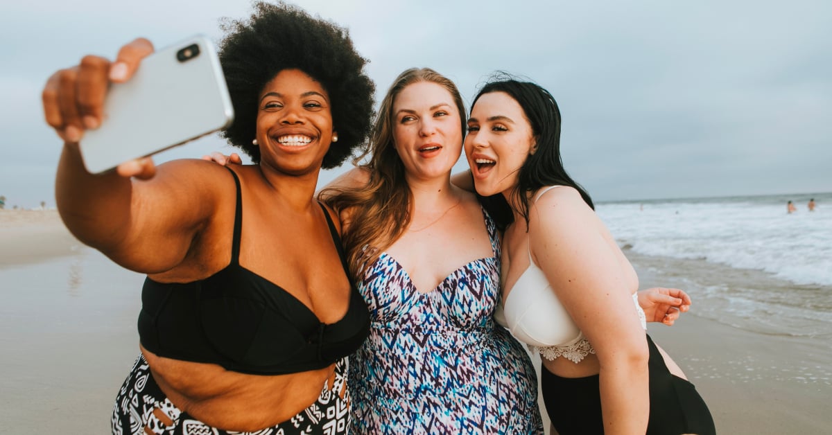 Three female-presenting people in swimwear pose in front of a phone on a beach to take a group selfie.